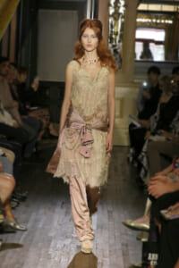 Model walking a fashion runway in a beige lace dress with a pink satin sash tied at the waist, surrounded by an audience.