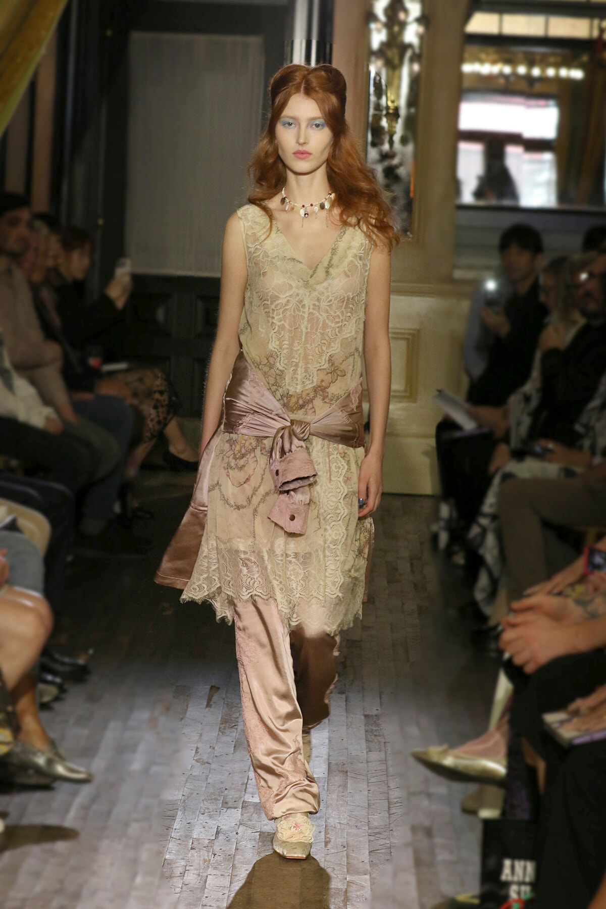 Model walking a fashion runway in a beige lace dress with a pink satin sash tied at the waist, surrounded by an audience.