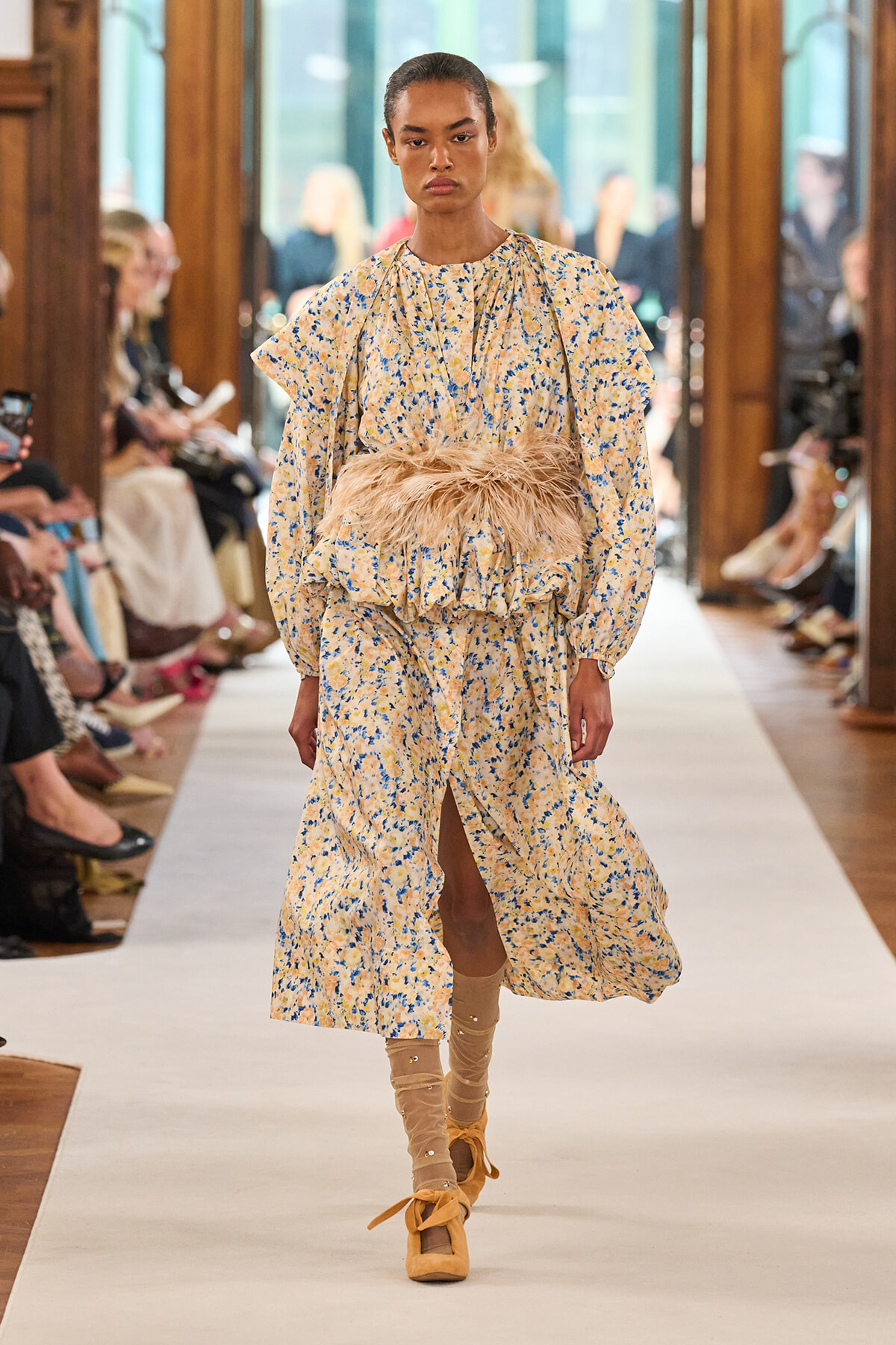 Model walking a fashion runway in a pale yellow floral dress with a feathered waist belt, audience on both sides watching.