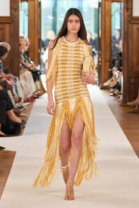 Model walks a fashion runway in a cream-and-yellow fringed dress with long yellow fringe skirt, holding a beige clutch.