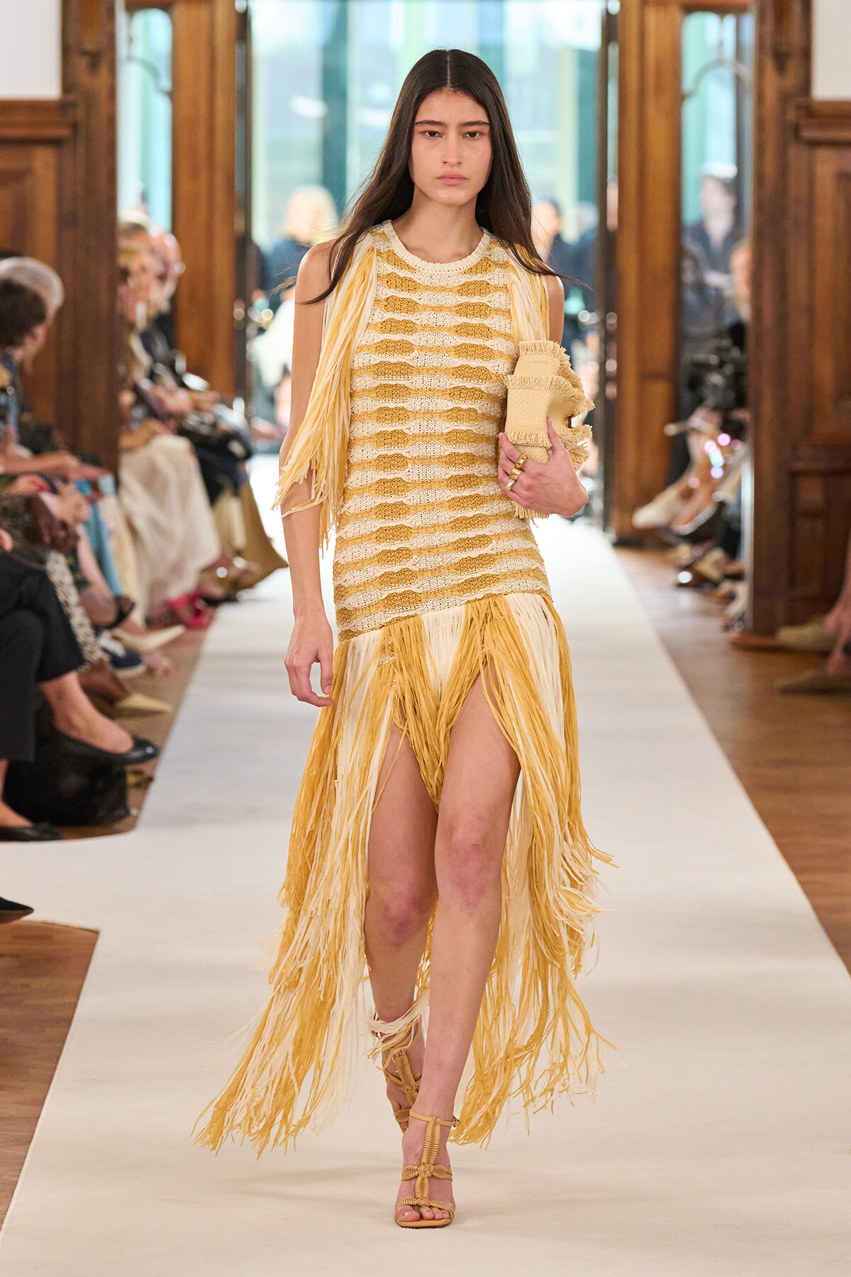 Model walks a fashion runway in a cream-and-yellow fringed dress with long yellow fringe skirt, holding a beige clutch.