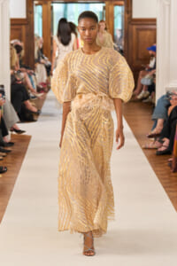 Model walks the runway in a gold, zebra-patterned dress with voluminous puff sleeves and a feather belt, paired with blue strappy heels. Audience seated along the catwalk.