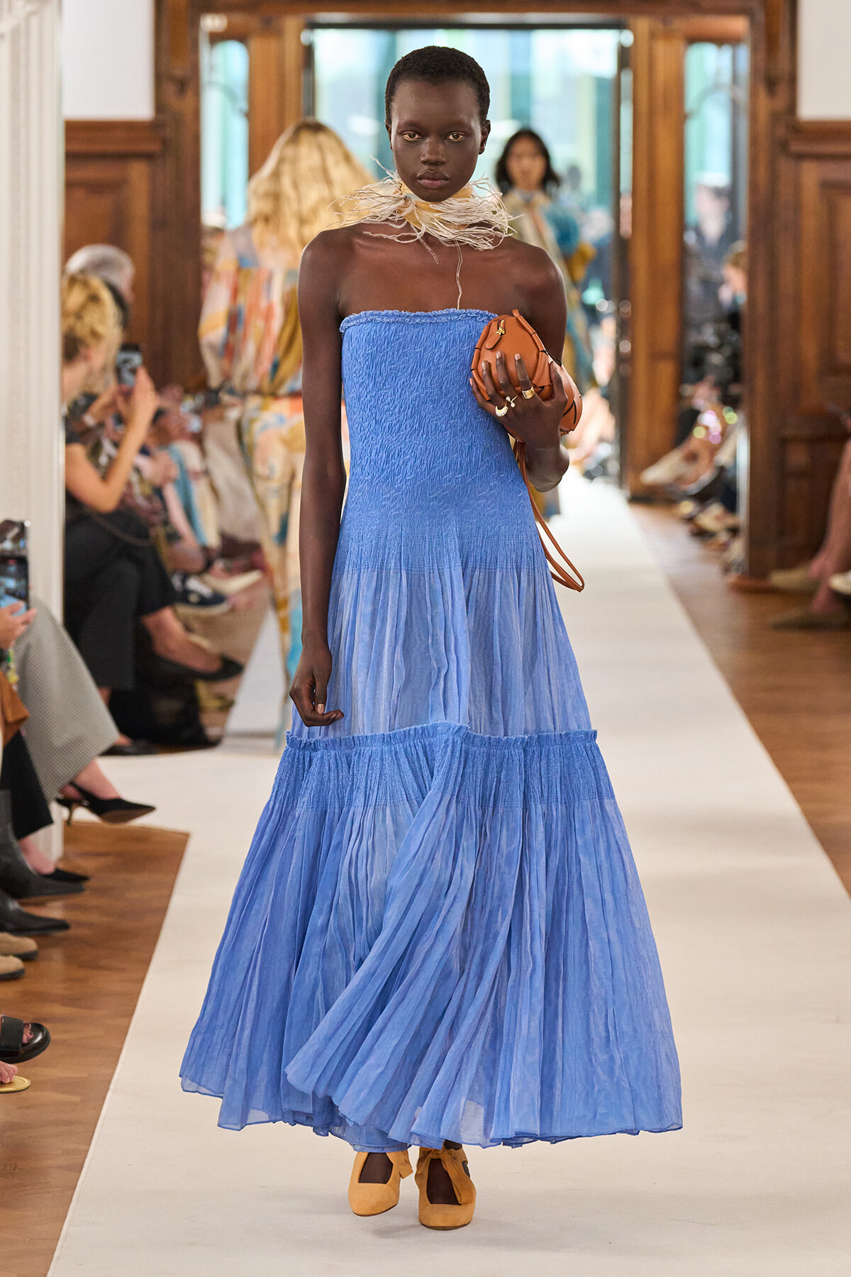 Black model walks runway in strapless light-blue tiered gown, holding tan clutch, beige feathered choker, and mustard heels.