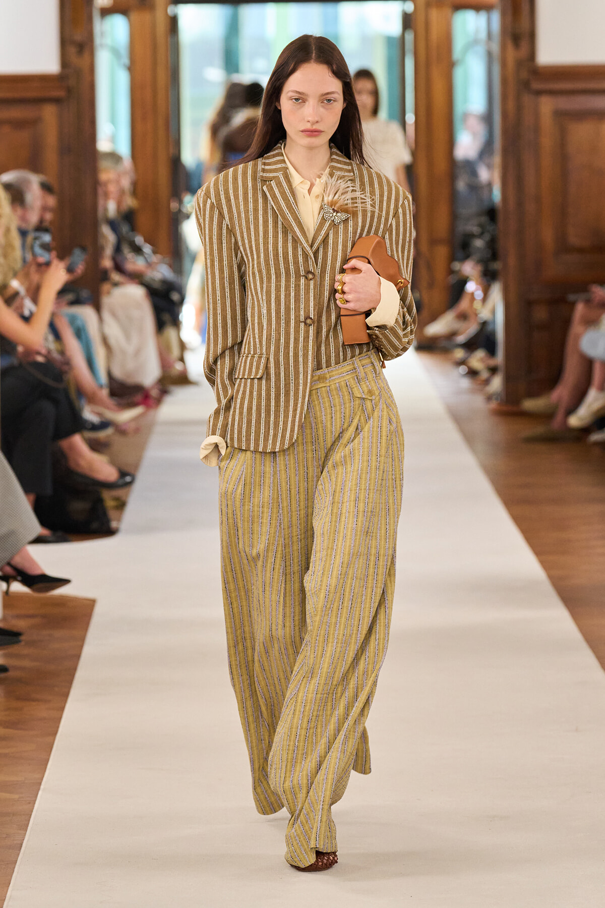 Model walks the runway in a tan striped blazer and matching wide-leg pants, holding a brown clutch, at a fashion show.