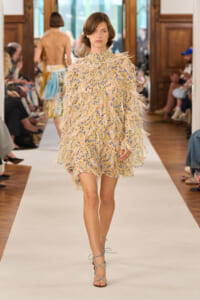 Model walks down a runway wearing a beige floral mini dress with feathered fringe and blue lace-up sandals during a fashion show.