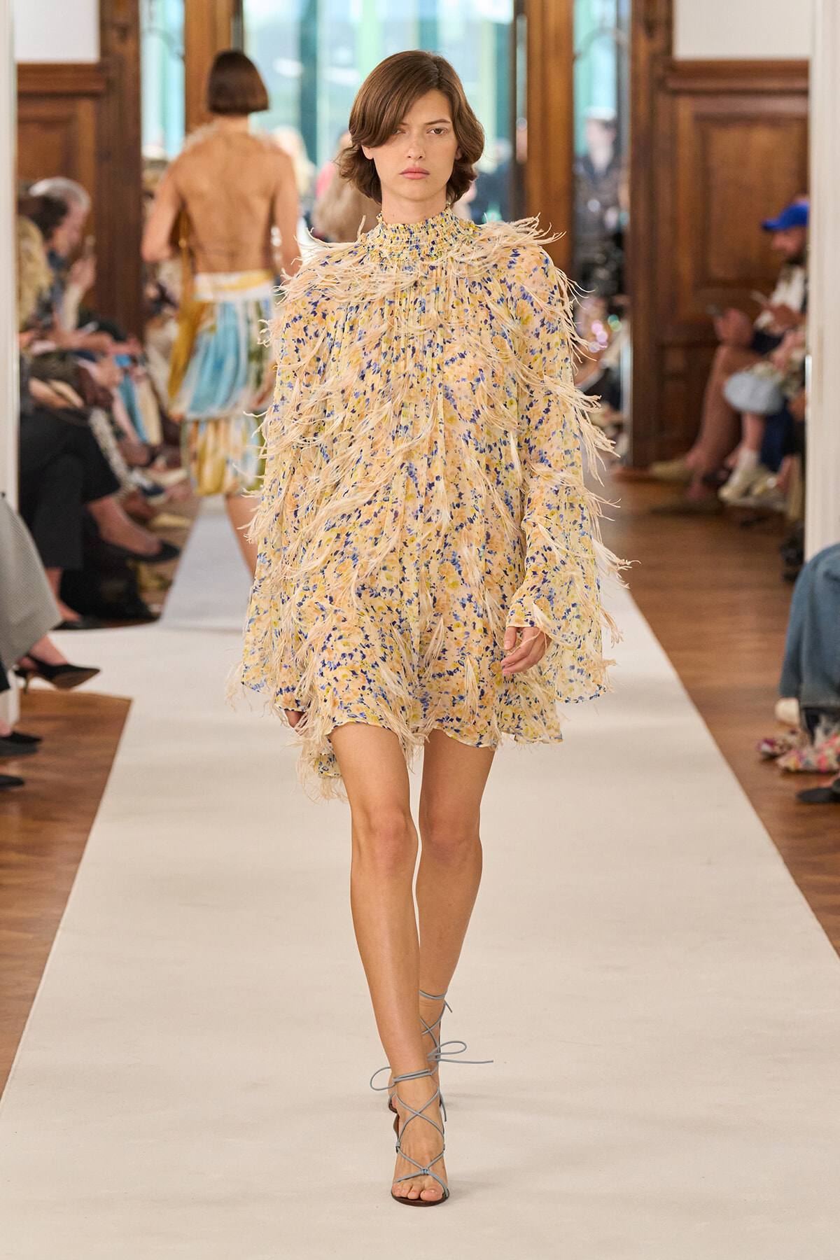 Model walks down a runway wearing a beige floral mini dress with feathered fringe and blue lace-up sandals during a fashion show.