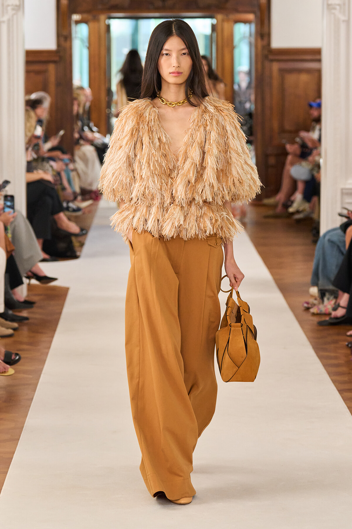 Model walking runway in a tan fringe blouse and wide-leg camel pants, carrying a matching tan handbag.