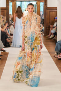 Model walking a runway in a long pastel tie-dye dress with beige feathered fringe embellishments on the sleeves and skirt.