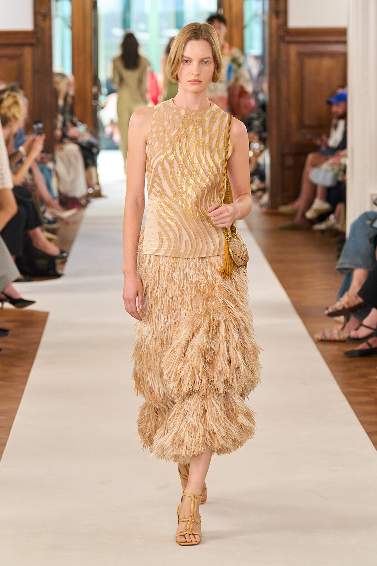 Female model walks a runway in a champagne-colored, sequined sleeveless top with a voluminous beige feathered skirt and gold sandals, carrying a small gold bag with tassels.