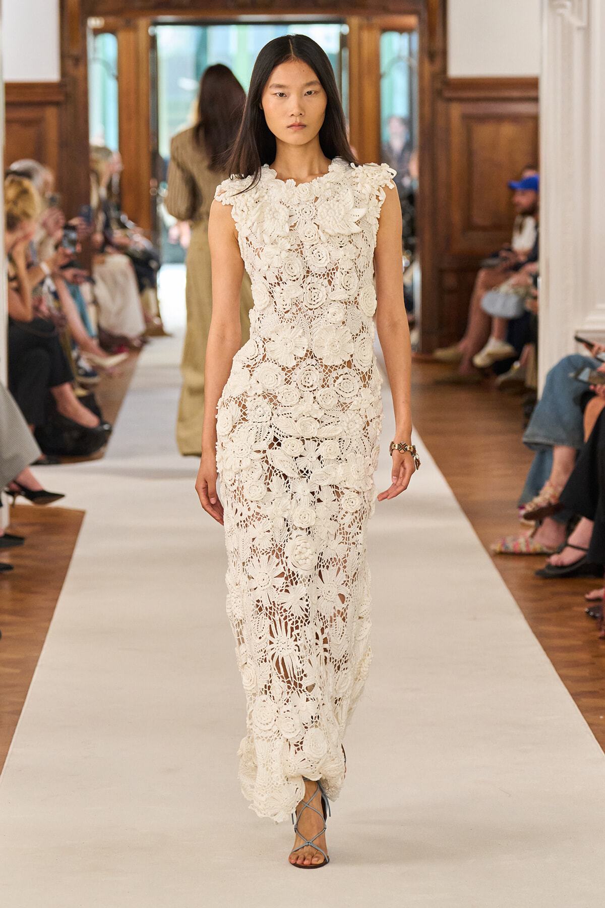 Model walks the runway in a full-length ivory lace dress with bold floral crochet patterns, paired with strappy sandals.
