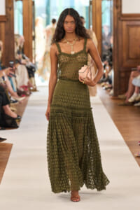 Model walks the runway in an olive green crochet mesh maxi dress, holding a pale pink clutch.
