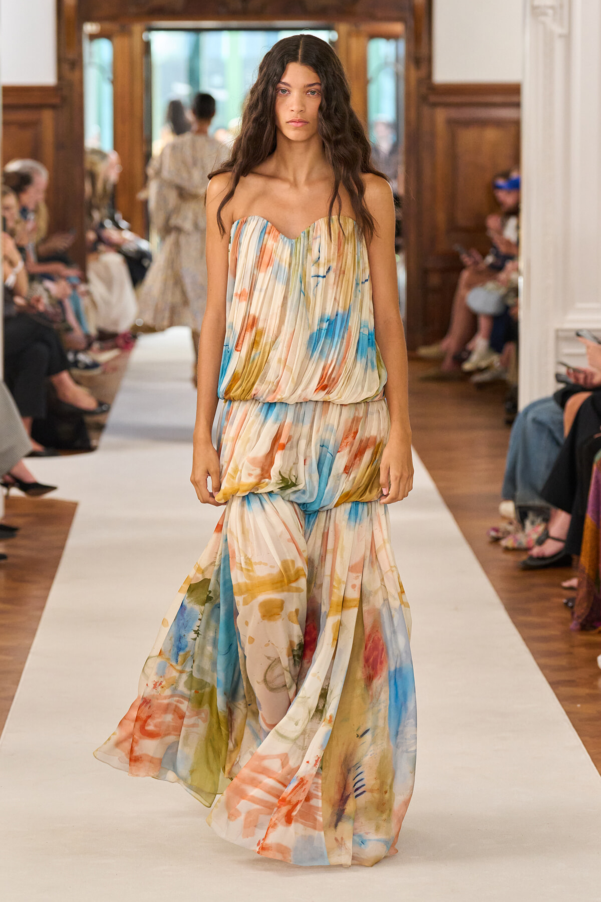 Model walks the runway in a strapless, pleated tie-dye maxi dress in pastel orange, blue, and green; audience watches from sides.
