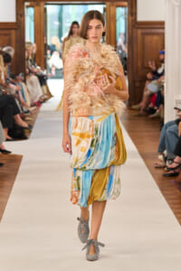 Model walks a runway in a fluffy pink-feathered top and multicolored draped skirt, holding a tan clutch and wearing gray bow-tied shoes.