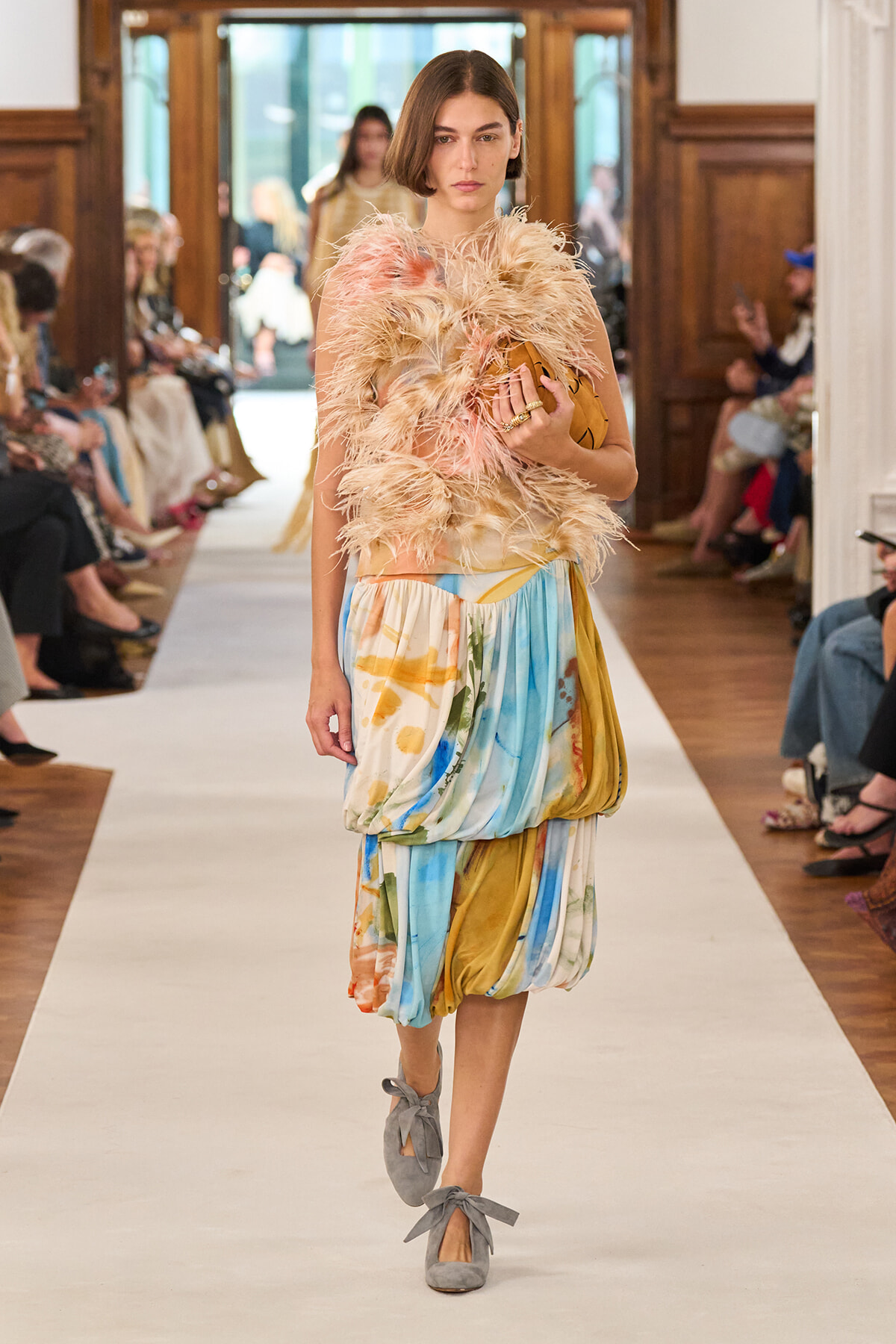 Model walks a runway in a fluffy pink-feathered top and multicolored draped skirt, holding a tan clutch and wearing gray bow-tied shoes.