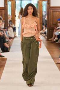 Model walking a runway in a peach ruffled sleeveless top and olive wide-leg trousers, carrying a peach fringe bag.