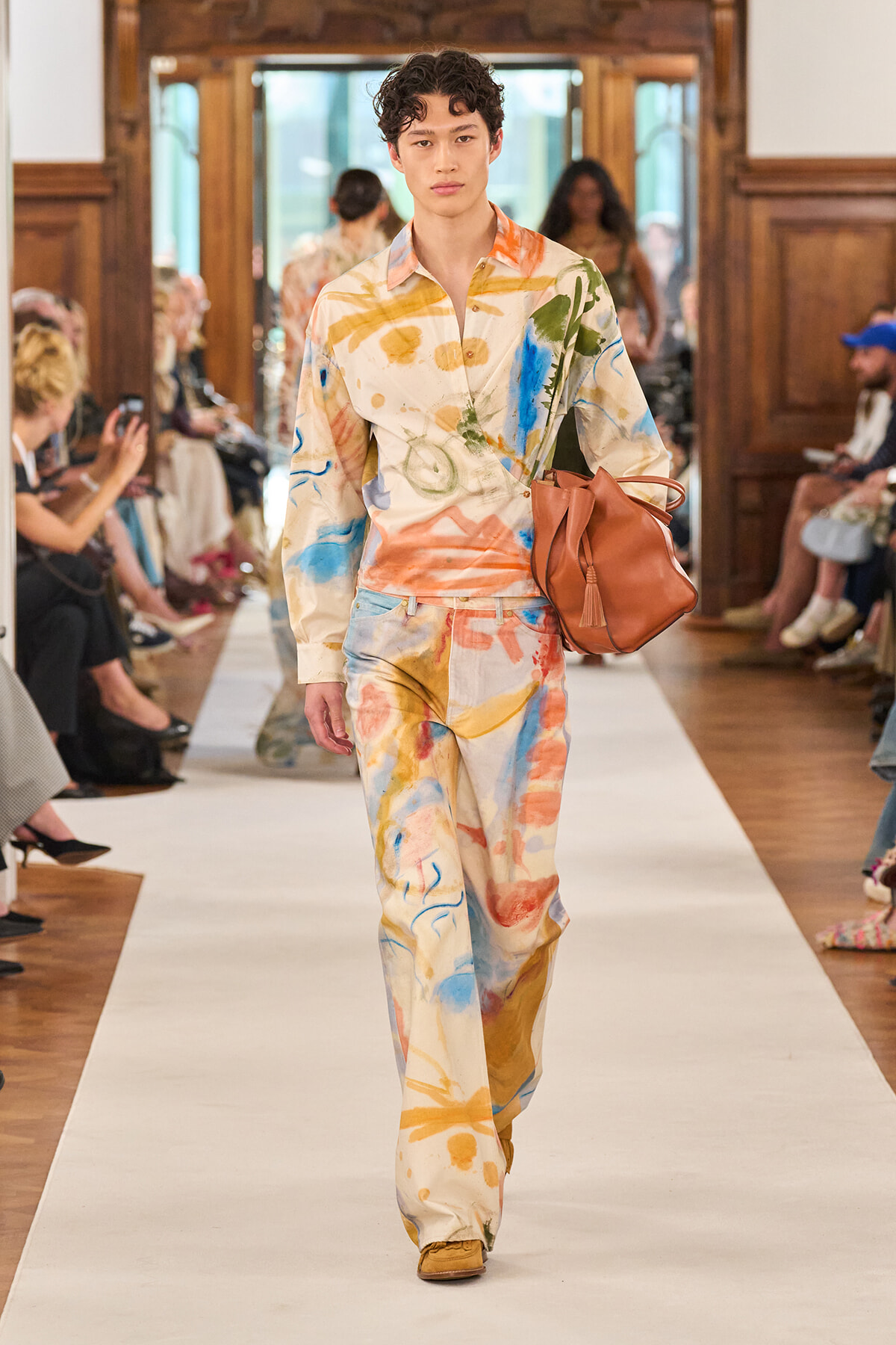 Male model walks a fashion show runway in a cream suit with multicolor paint-splatter print, carrying a brown leather tote bag.