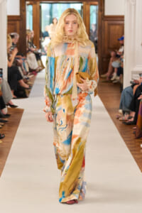 Model walking a runway in a long, multicolored abstract-print jumpsuit with a tied neck scarf, carrying a tan clutch.