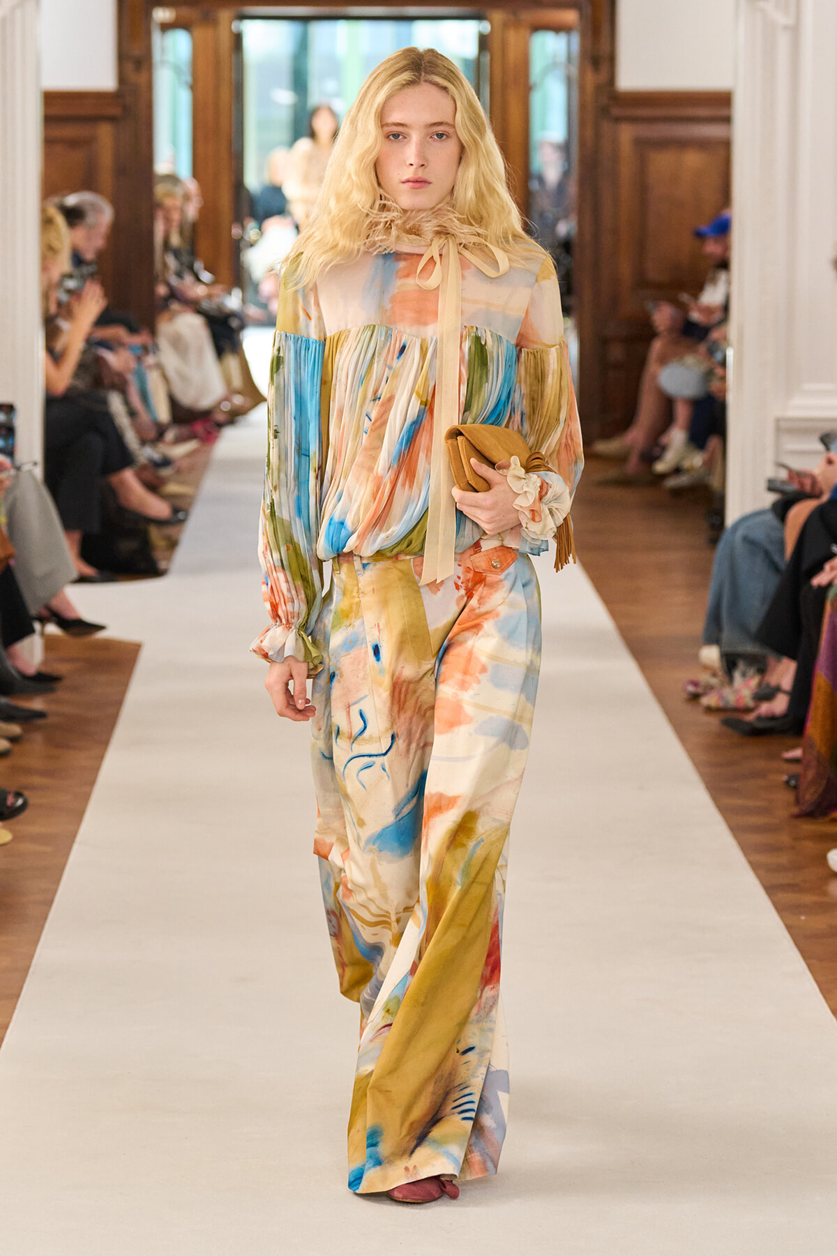 Model walking a runway in a long, multicolored abstract-print jumpsuit with a tied neck scarf, carrying a tan clutch.