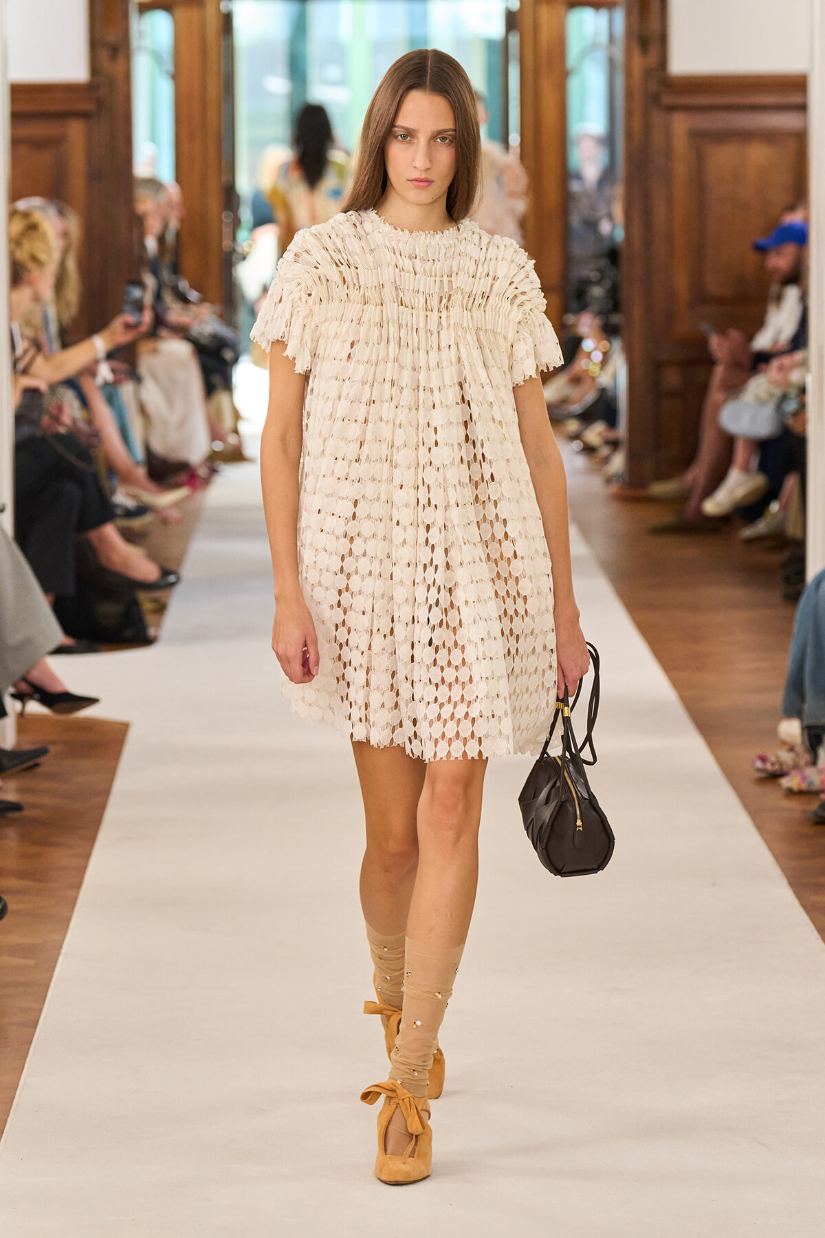 Model walks a runway in a cream crochet dress, beige knee-high socks with bows, and tan platform shoes, carrying a black handbag.
