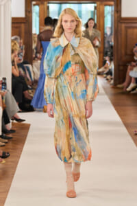Model walks the runway wearing a long, pastel watercolor-print dress with voluminous puff sleeves and a gathered waist, orange sandals visible.