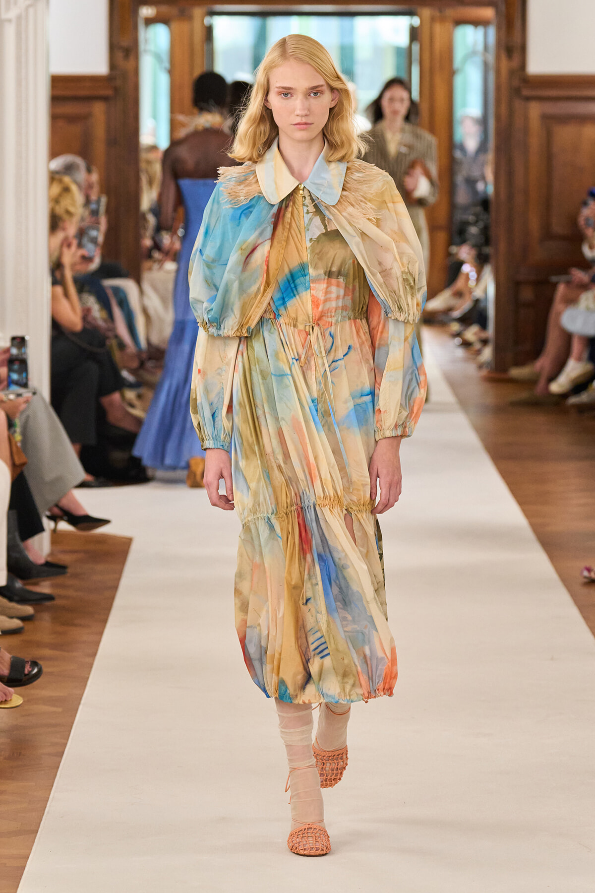 Model walks the runway wearing a long, pastel watercolor-print dress with voluminous puff sleeves and a gathered waist, orange sandals visible.