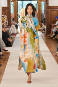 Model walks a fashion runway in a loose, abstract-print maxi dress in blue, green, orange, and yellow, with a scarf-like drape and brown lace-up flats.