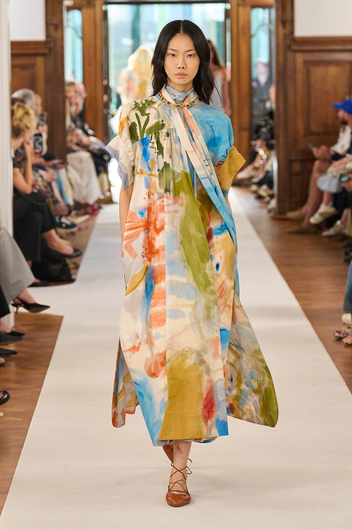 Model walks a fashion runway in a loose, abstract-print maxi dress in blue, green, orange, and yellow, with a scarf-like drape and brown lace-up flats.