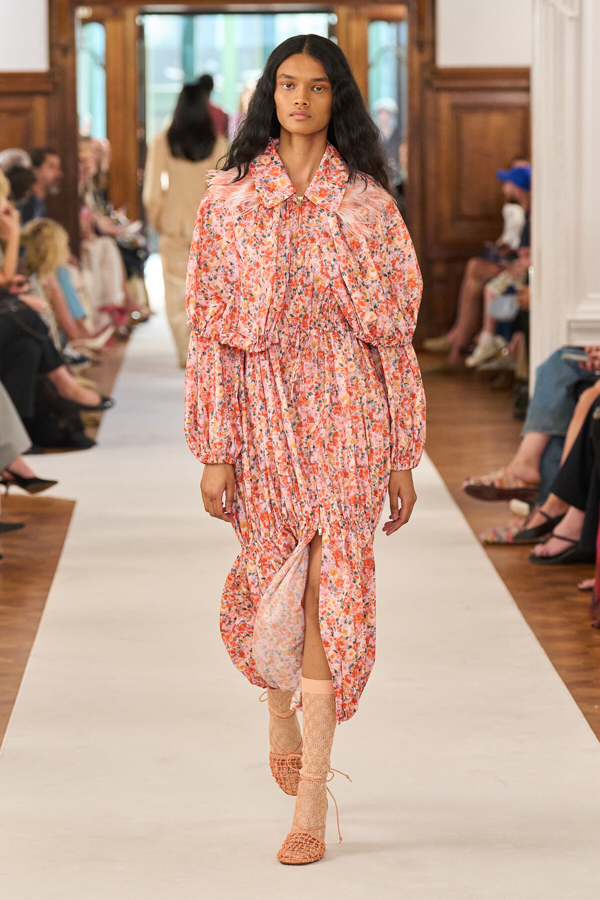 Model walking the runway in a pink floral print maxi dress with puff sleeves and a front slit, paired with peach lace-up sandals.