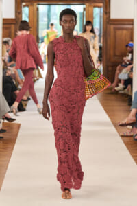 Female model on a runway wearing a pink lace floor-length dress, carrying a multicolor woven tote bag on her shoulder.
