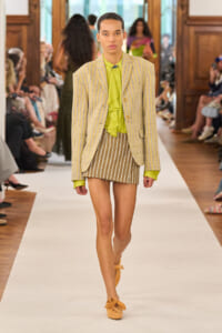 Model on a runway wearing a lime green blouse with a ruffled front, a beige striped blazer, a matching striped mini skirt, and orange bow-tied flats.