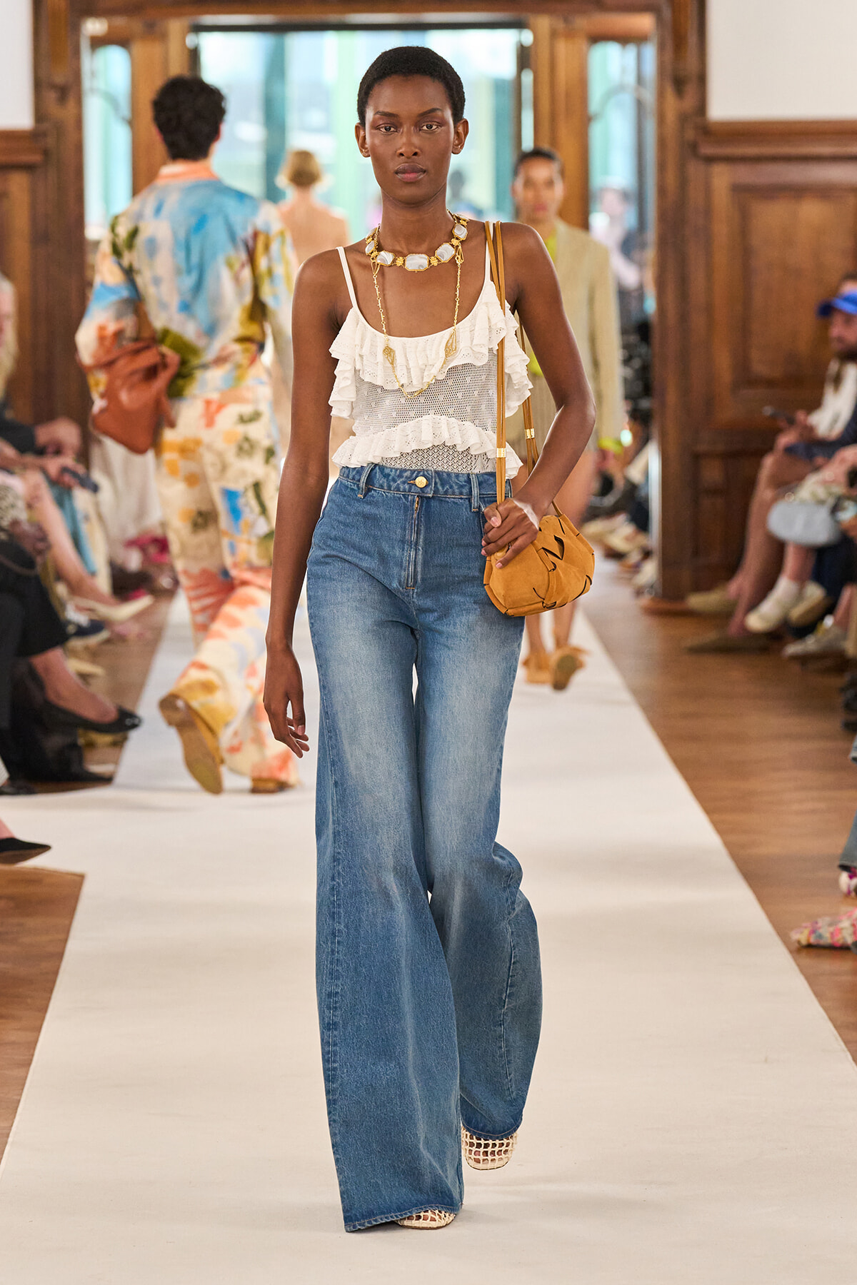 Model on a runway wearing a white ruffled sleeveless top, wide-leg blue jeans, tan crossbody bag, and layered gold jewelry.