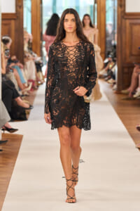 Model walking a fashion show in a black sheer lace dress with an intricate lattice pattern and strappy heels.