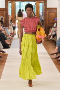 Model walking a runway in a pink textured crop top and lime green tiered maxi skirt, carrying a colorful orange bag.
