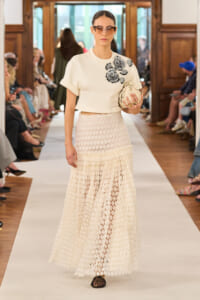 Model walks a runway in a white cropped top with floral embroidery and a cream lattice maxi skirt, carrying a small clutch and wearing sunglasses.