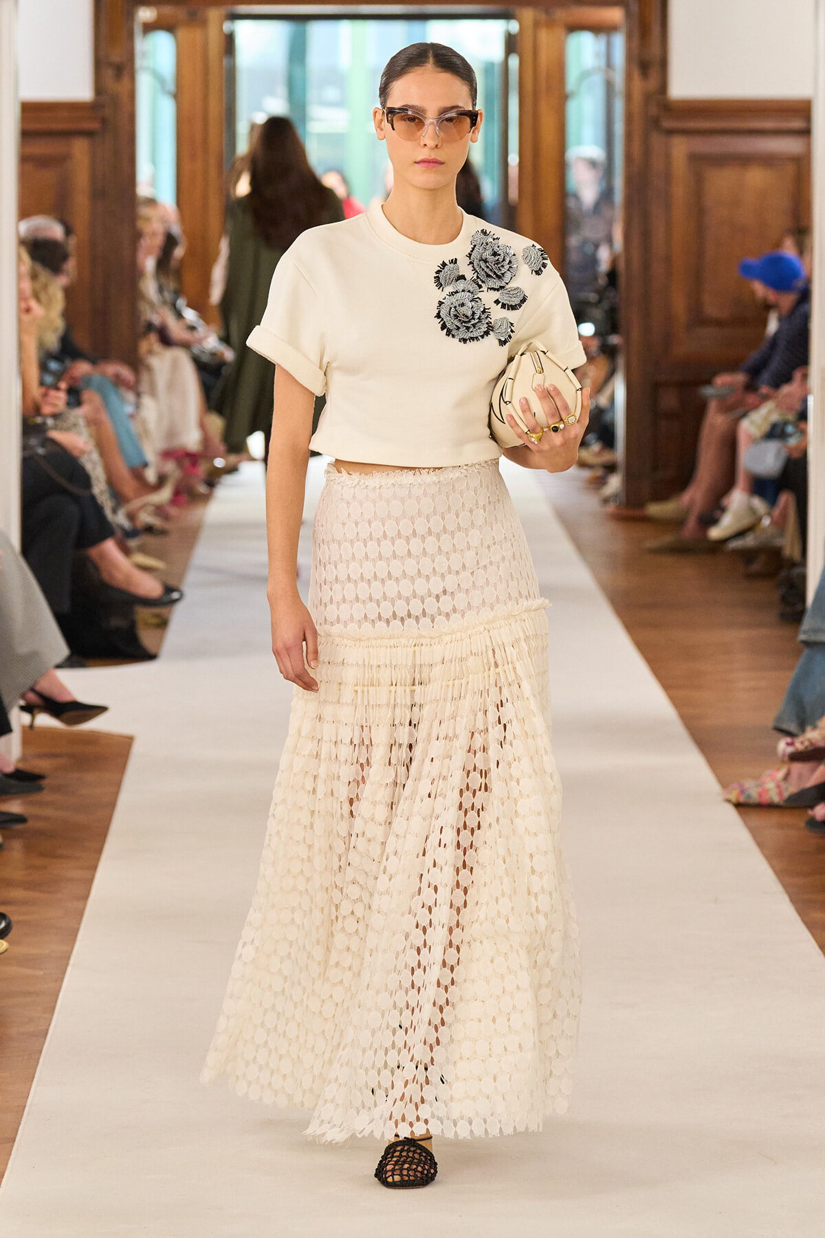 Model walks a runway in a white cropped top with floral embroidery and a cream lattice maxi skirt, carrying a small clutch and wearing sunglasses.