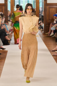 Model walking a fashion show wearing a beige textured sleeveless top, a tan smocked long skirt, yellow sunglasses, and carrying a cream ruffled clutch.
