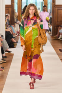 Model walking a fashion show wearing a long, multicolored abstract-print coat and carrying a brown suede tote bag; pink shoes visible along the white runway.