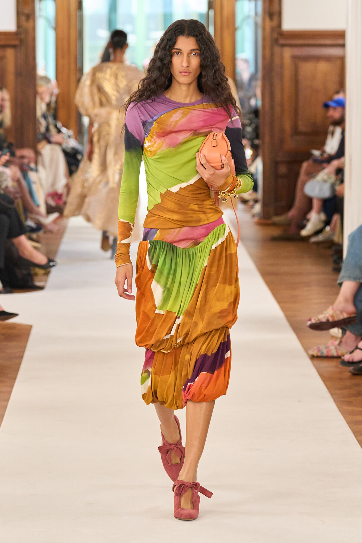 Model walking the runway in a vibrant color-blocked, long-sleeve dress with pink ankle-strap shoes, holding a peach clutch.
