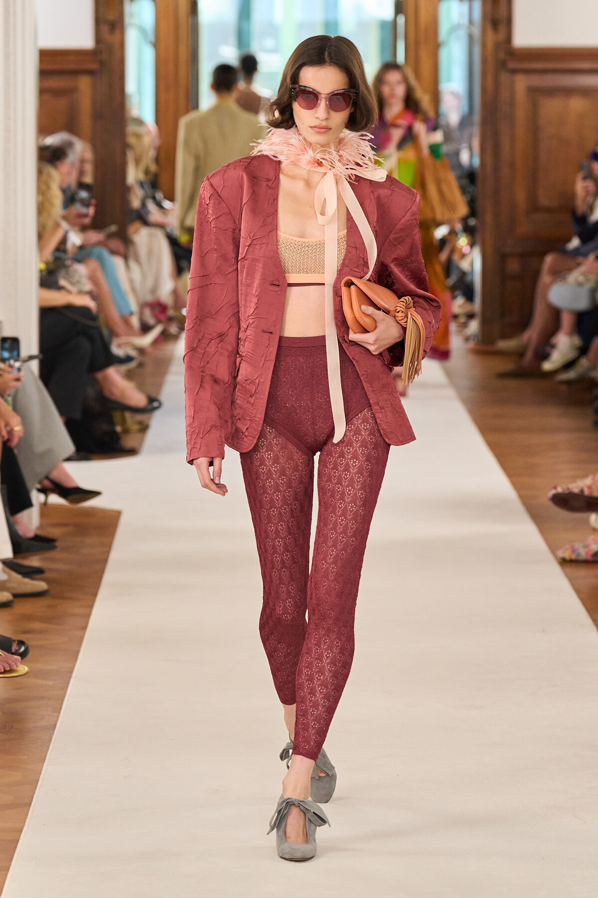 Model wearing a mauve textured blazer with matching patterned burgundy tights, pink sunglasses, feather neck scarf, and gray bow-strap heels on a runway.