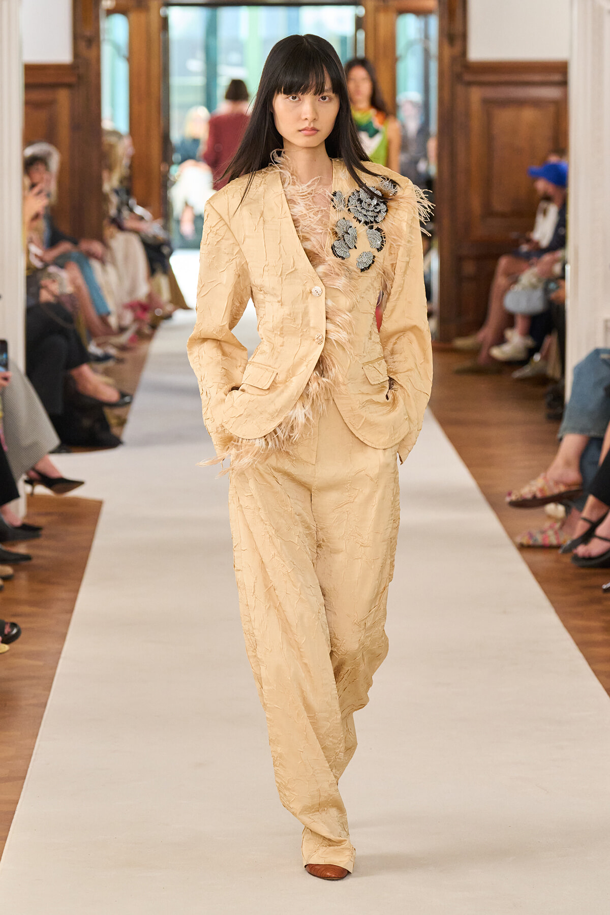 Model walks the runway in a beige textured pantsuit with feathered fringe and a blue-gray floral appliqué on the left shoulder.