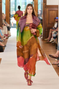Model walks a runway in a sheer, colorful layered outfit, holding a pink textured scarf.