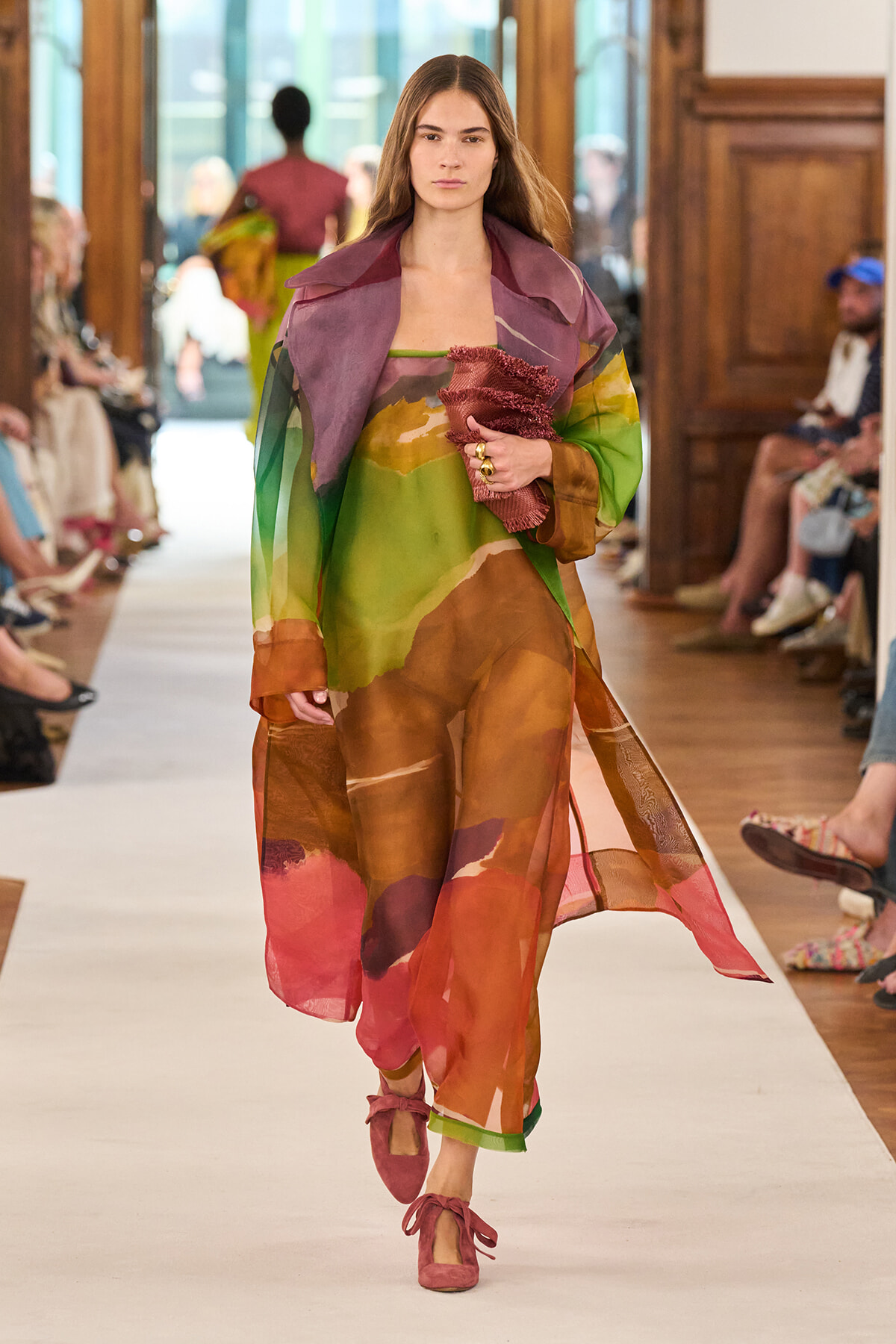 Model walks a runway in a sheer, colorful layered outfit, holding a pink textured scarf.
