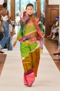 Model walking a runway in a sheer, multicolored abstract-print blouse paired with matching wide-leg pants and a chunky gold necklace, audience in background.