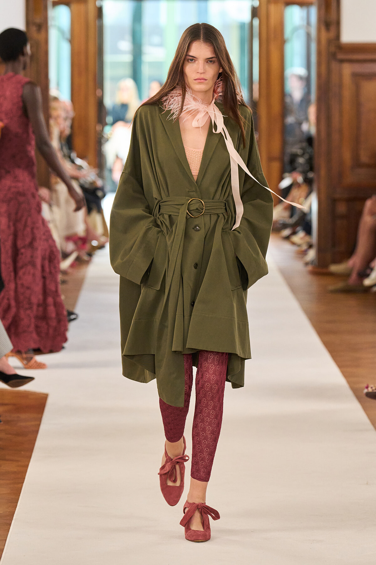 Model walking a runway in an oversized olive trench with belt, pale pink neck scarf, burgundy patterned leggings, and pink bow-tied flats.