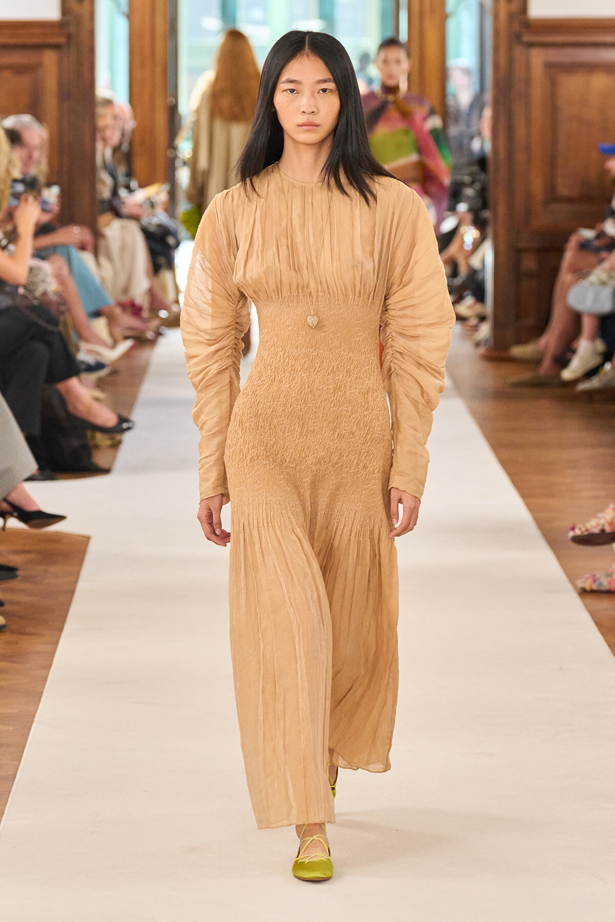 Model walking the runway in a beige textured long-sleeve maxi dress with a smocked waist; audience seated on both sides of the catwalk.
