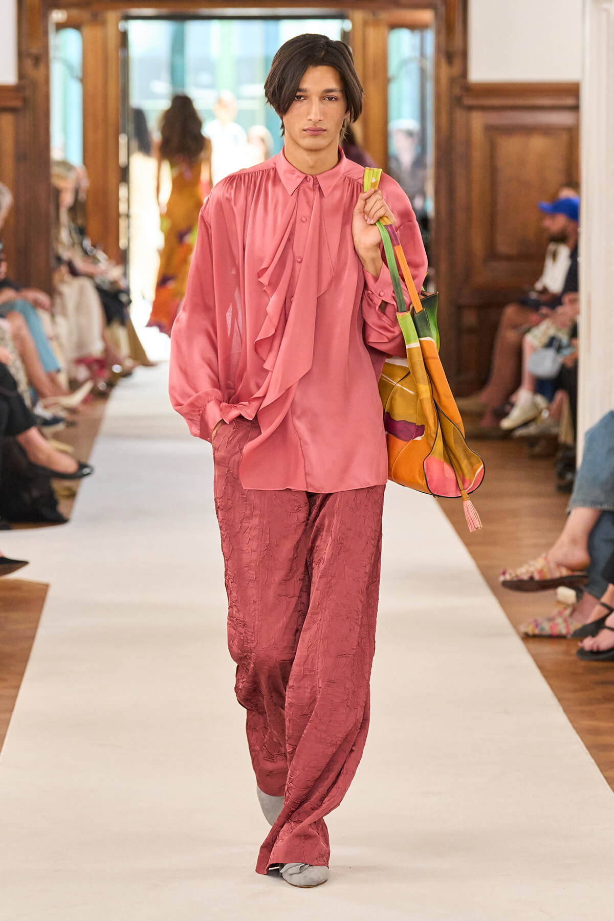 Model wearing a pink satin blouse with ruffles and matching loose pants, carrying a multicolored tote bag down a fashion runway.