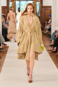 Model walks a fashion runway in a beige belted trench coat, pink patterned tights, and brown lace-up heels; feathered neck accessory visible.