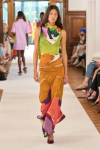 Model walking a runway in a lime-green sleeveless top, multicolored scarf, and a flowing abstract orange–pink dress.