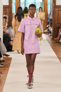 Model walking a fashion show in a lavender utility dress with gold buttons, yellow sunglasses, a neon yellow handbag, and gray bow-strap shoes with burgundy socks.