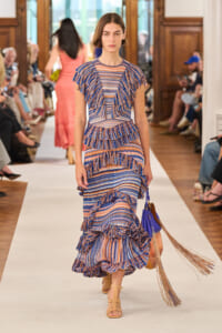 Model walking the runway in a blue-and-orange striped, layered ruffle dress with a purple handbag and tan sandals; audience seated along the catwalk in a fashion show venue.